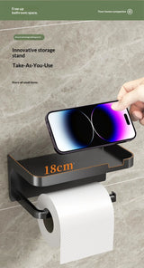 Black wall-mounted toilet paper holder with a roll of white toilet paper, shown against a gray marble-look wall. A hand is holding a smartphone above the holder, which also has a phone placed on its top surface.